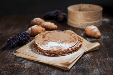 freshly baked sweet pancakes stacked on brown baking paper, croissants and grapes on a dark brown wooden table on a dark gray background