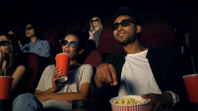 Cheerful multiethnic hipsters talking and smiling while watching film and holding cola and popcorn on cinema
