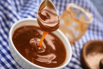 Spoon with tasty caramel sauce over bowl, closeup
