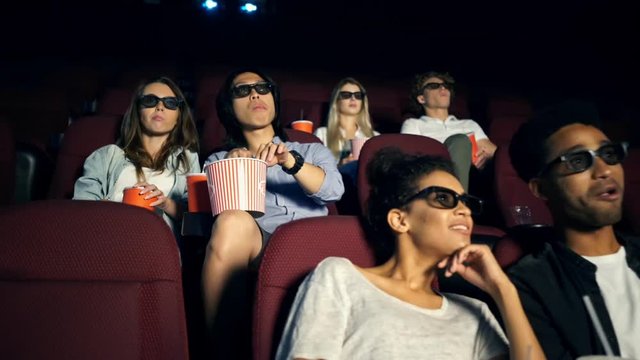 Young concentrated multiethnic audience friends watching film with popcorn and talking in cinema