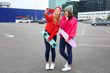 Two happy hipster girls with skateboards taking selfie outdoors
