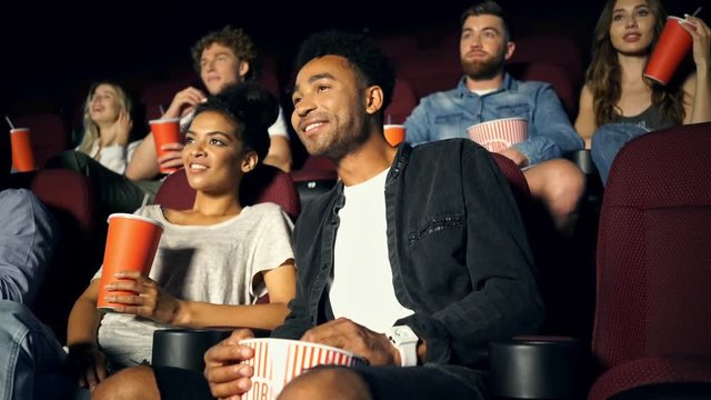 Young positive multiethnic spectators watching film with popcorn and smiling in cinema