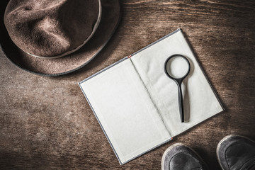 Opened old blank book with magnifying glass. Detective hat and shoes on brown background. Retro things. Historical atmosphere. Vintage english style. Evidence searching concept. Empty place for text.