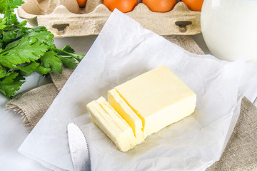 A bar of butter is cut into pieces on a wooden board with a knife, surrounded by milk, eggs and parsley on a white table. Ingredients for cooking.