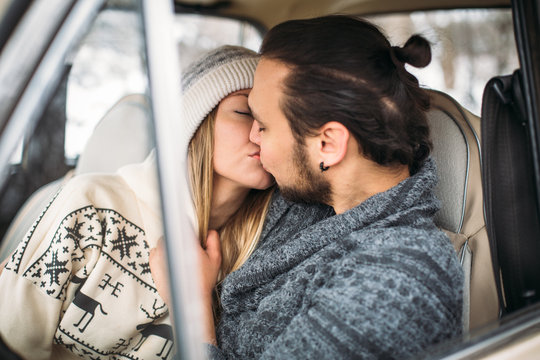 A Valentine couple sitting inside a snow retro car and kissing in a forest. Romantic Beauty girl in a hat and handsome man in a grey pullover. Horisontal picture