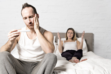 Sad Man Sitting On Bed With pregnant woman behind look at pregnant test