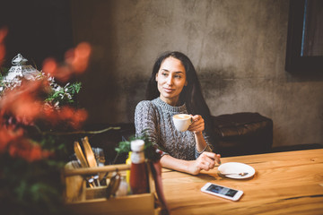 beautiful young girl drinks coffee from a white cup, next to her cell phone in a cafe decorated...
