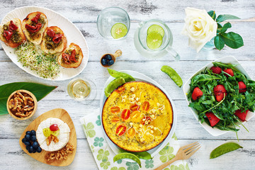 Frittata (omelet) with potato, tomato and pea sprouts, bruchetta, cheese plate, salad with strawberry and ruccola and lemonade. Top view of brunch or lunch on a white table.