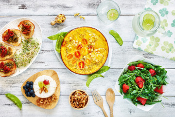 Frittata (omelet) with potato, tomato and pea sprouts, bruchetta, cheese plate, salad with strawberry and ruccola and lemonade. Top view of brunch or lunch on a white table.