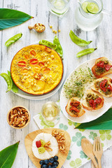 Vertical top view of healthy lunch or dinner on white table. Frittata with young peas, bruchetta with tomato and pesto, cheese plate and lemonade. 