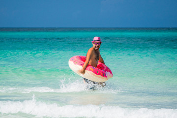 Happy handsome man in pink hat runs with a big inflatable tube on the coast of Caribbean Sea in summer sunny day