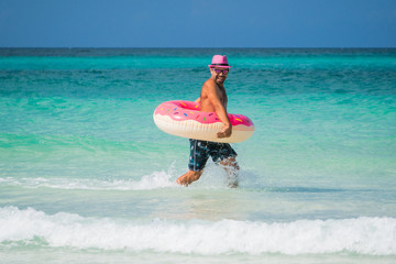 Naklejka na ściany i meble Happy handsome smiling man in pink hat runs with a big inflatable tube on the coast of Caribbean Sea in summer sunny day