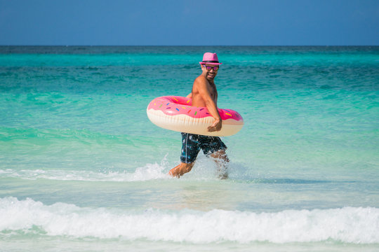 Happy handsome smiling man in pink hat runs with a big inflatable tube on the coast of Caribbean Sea in summer sunny day