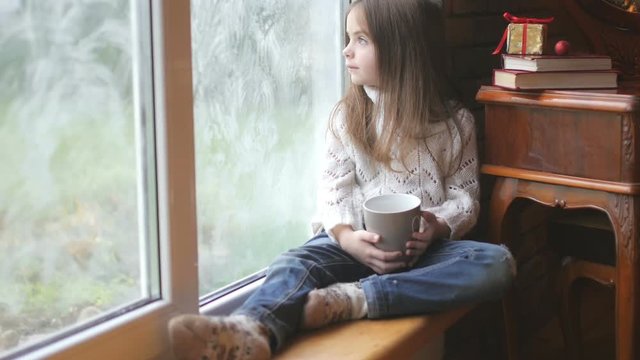 beautiful little girl with a mug of hot tea sits by the window, looks at the street and what she dreams about