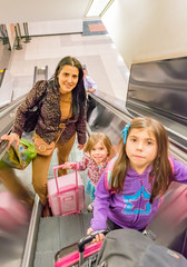 European pretty young mom and her two daughters and their luggage go up an escalator in the airport. 