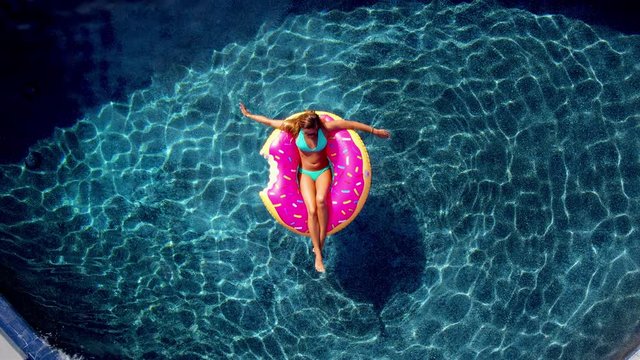 Young happy woman relaxing on inflatable pool toy in blue swimming pool on sunny day waiving at camera 