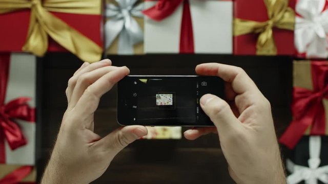 Adult man takes photo of beautiful Xmas present on smartphone camera on decorated holiday desk, top down shot