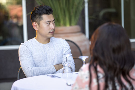 Handsome Asian Man On An Outdoor Date With A Black Female.  The Couple Are Sitting In A Restaurant Or Cafe Setup For Speed Dating.  He Looks Bored And Disappointed.