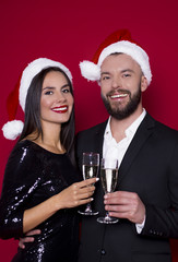 Have a nice new year and christmas. Cheerful beautiful smiling couple of lovers in Santa hats with the glasses of champagne on a red background with balls. Lovers on holiday