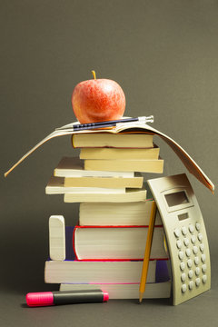 Concept of education. Stack of school books with calculator, pink marker and pencil and a red apple on top in front of dark gray background