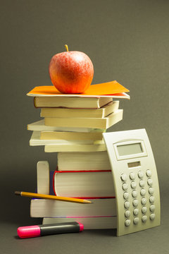 Concept of education. Stack of school books with calculator, pink marker and pencil and a red apple on top in front of dark gray background