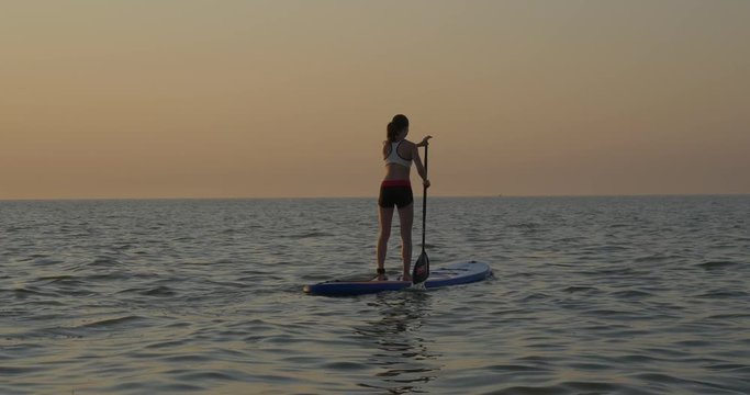 Athletic girl rowing on standup paddle board in the evening on the blue water