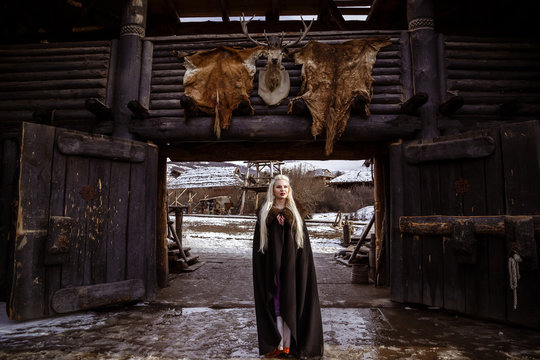 Outdoors portrait of beautiful furious scandinavian warrior ginger woman in a traditional clothes with fur collar, with sword in her hand and wooden Viking Village view on the background.