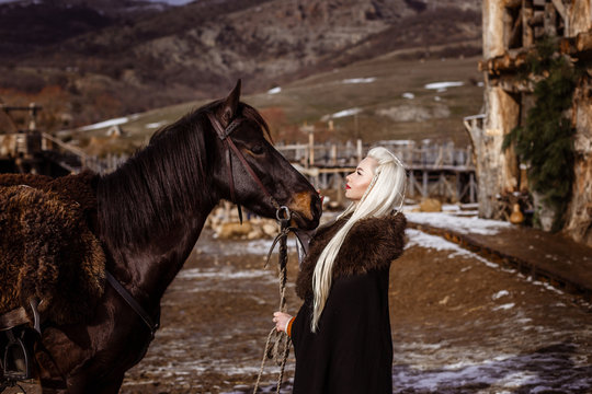 Outdoors portrait of beautiful furious scandinavian warrior ginger woman in a traditional clothes with fur collar, with sword in her hand and wooden Viking Village view on the background.