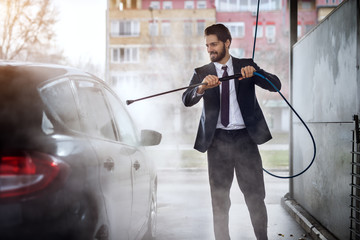 Happy cheerful young stylish businessman in suit washing his car at the manual car washing self-service station.