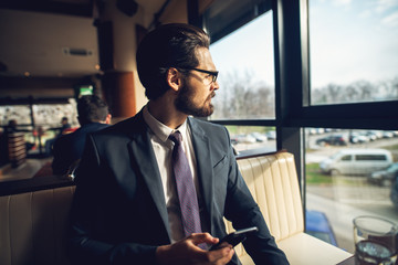 Close up side view of stylish bearded handsome young businessman in suit sitting at a cafe upstairs and looking far away while holding a mobile.