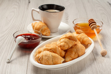 Cup of black coffee with saucer, fresh croissants on white porcelain dish, honey and strawberry jam in bowls