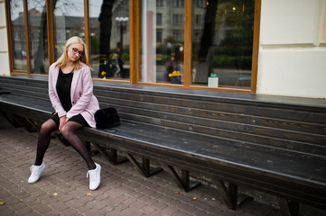 Blonde girl at glasses and pink coat, black tunic sitting on bench at street.