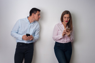 Middle-aged couple with smartphones. Husband in casual clothes peeking into his pretty wife's smartphone which typing message to her lover on white background