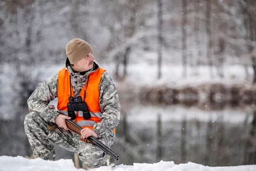 Photo sur Plexiglas Chasser Male hunter in camouflage, armed with a rifle, sittiing in a snowy winter forest