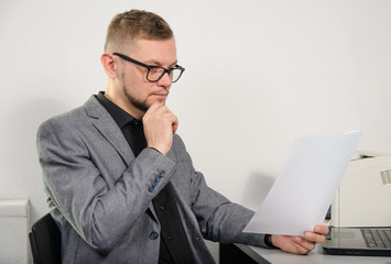 a man in glasses and a jacket thoughtfully studying the documents for work at the table with a laptop and a printer in the office