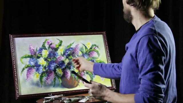A man bearded artist in a blue sweater draws an artistic brush painting flowers still life on easer in the studio
