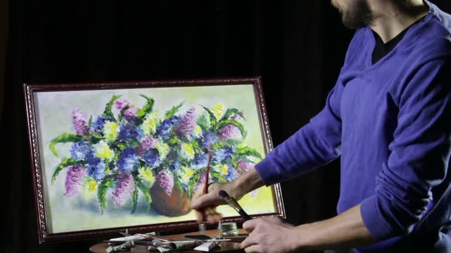 A man bearded artist in a blue sweater draws an artistic brush painting flowers still life on easer in the studio