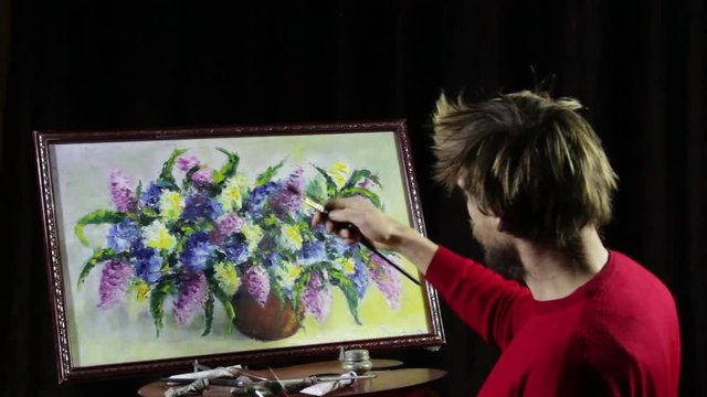 A man bearded artist in a red sweater draws an artistic brush painting flowers still life on easer in the studio