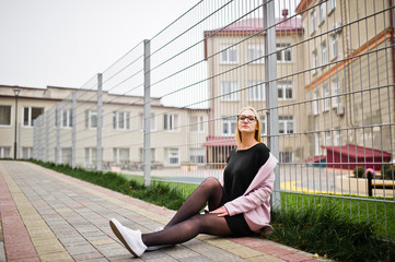 Fototapeta na wymiar Blonde girl at glasses and pink coat, black tunic sitting against fence at street.