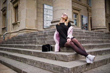 Blonde girl at glasses and pink coat with handbag sitting on stairs of opera building.