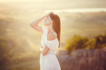 Portrait of brunette attractive woman with light smile and closed eyes for enjoy moment of walking alone in wild rocks on sunset. Girl wears white retro dress