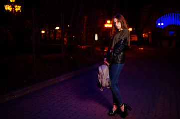 Night portrait of girl model wear on glasses, jeans and leather jacket, with backpack in hands, against blue lights garland of city street.