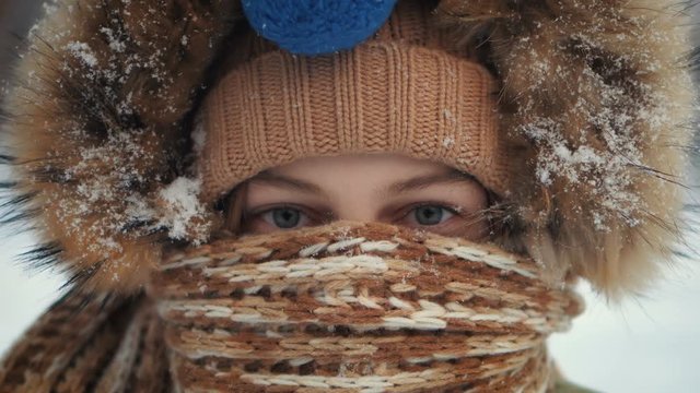 Cute close-up portrait of a young girl covered her face with knitted hat and scarf. Snowy weather. Winter time. Real people series