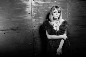 Studio portrait of sexy blonde girl with originally make up on neck, wear on luxurious fur coat at gray iron steel background.