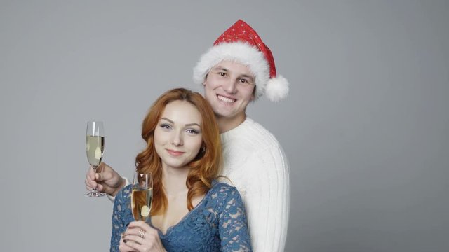 Young couple with glasses of champagne, man wearing Santa hat, looking at camera and lifting glasses cheering