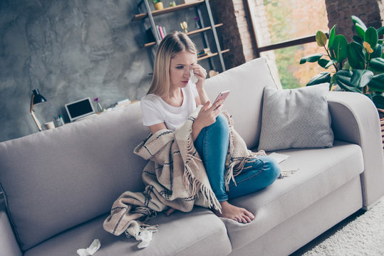 Single frustrated exhausted woman is sitting on a sofa in a living room covered with a plaid, surrounded with paper tissues, she is crying and reading old sms on her telephone