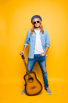 Vertical full length portrait of professional, successful musician in hat and glasses holding hand in pocket, stand guitar on the floor over yellow background
