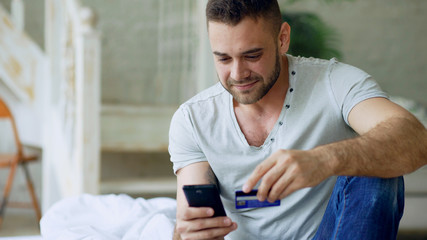 Attractive young man with smartphone and credit card shopping on the internet sit on bed at home