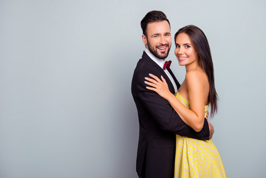 Portrait photo of fashionable sexy woman in yellow dress and bearded brunette man in black suit with red bow tie, wife and husband hugging, looking at camera, standing over grey background
