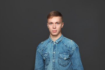 Obraz premium Portrait of young handsome man in jeans shirt looking at camera over dark grey background. Emotions and facial expressions concept.